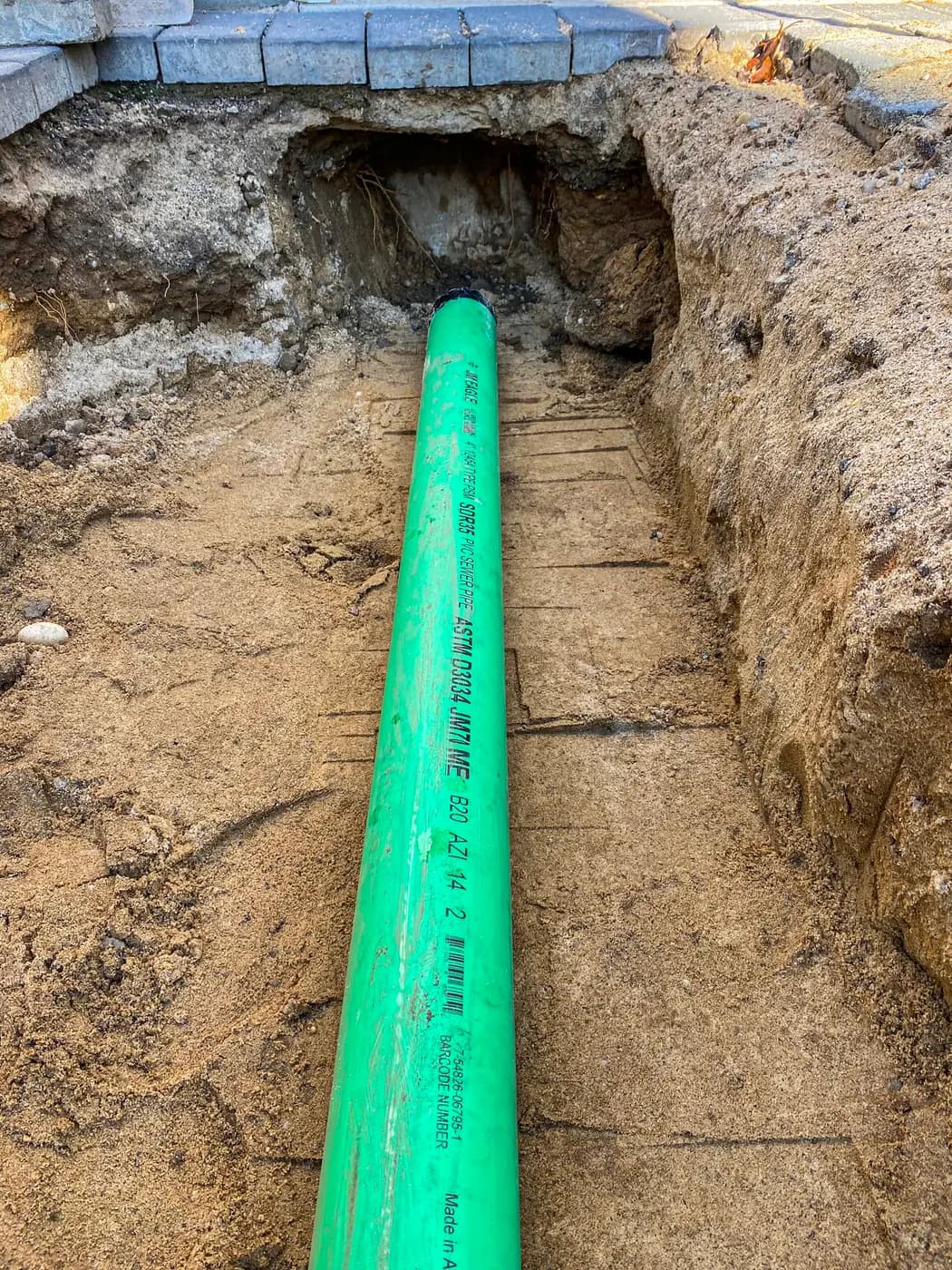 Green PVC sewer main laid in a trench beside a paver walk