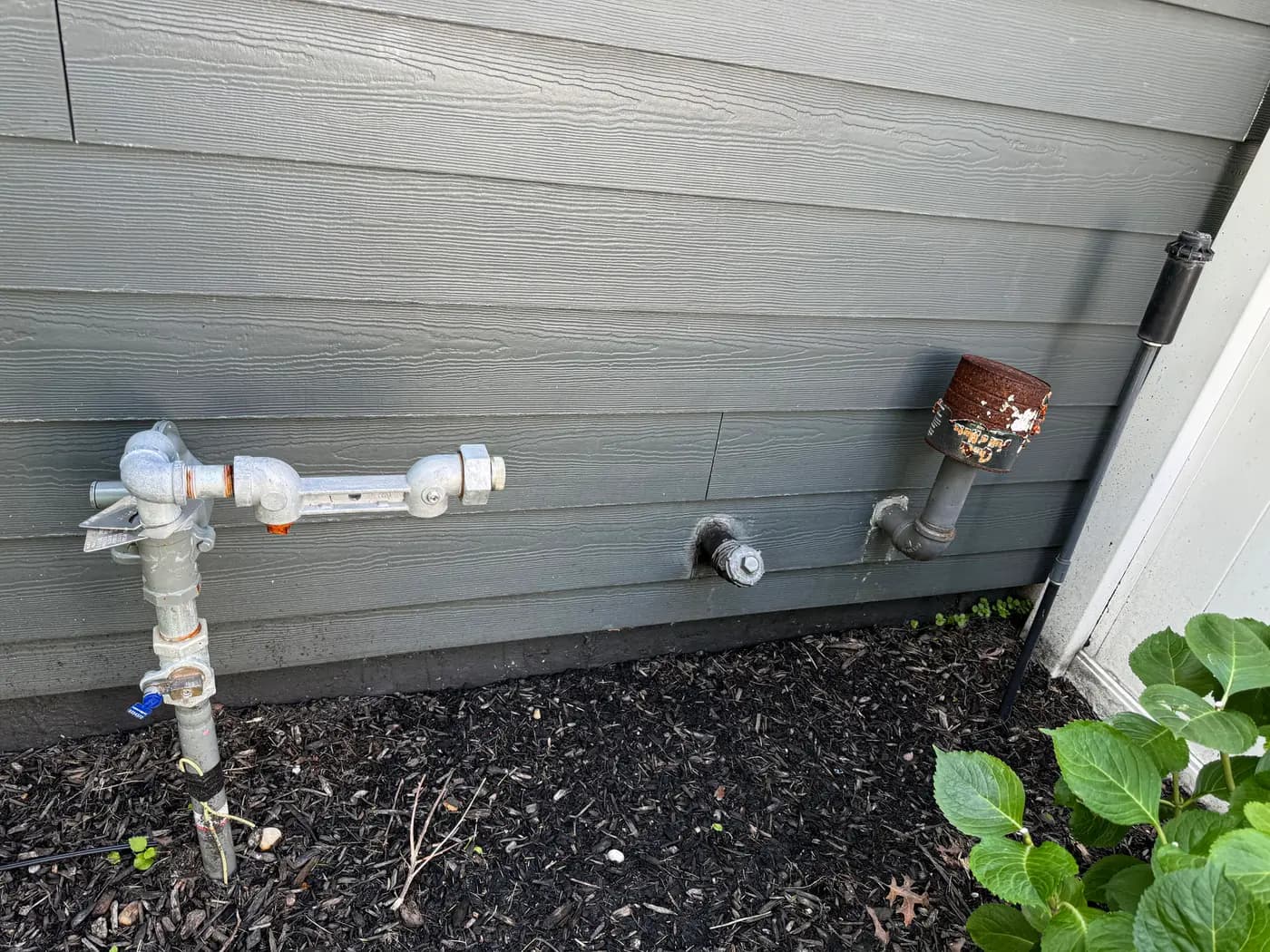 Galvanized gas riser with shutoff valve against gray siding above mulch bed