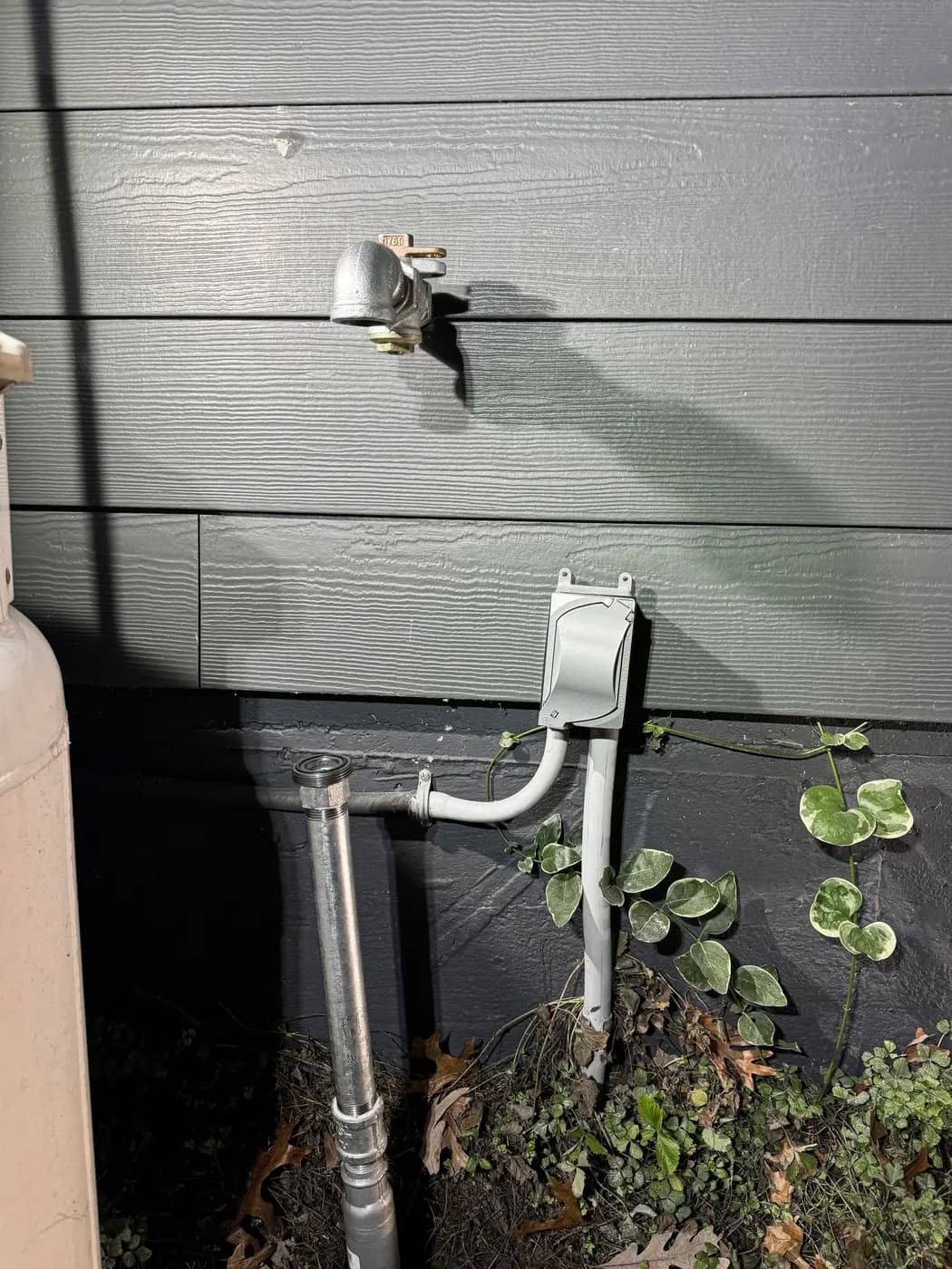 Galvanized riser and vented fitting mounted on gray siding above ivy groundcover