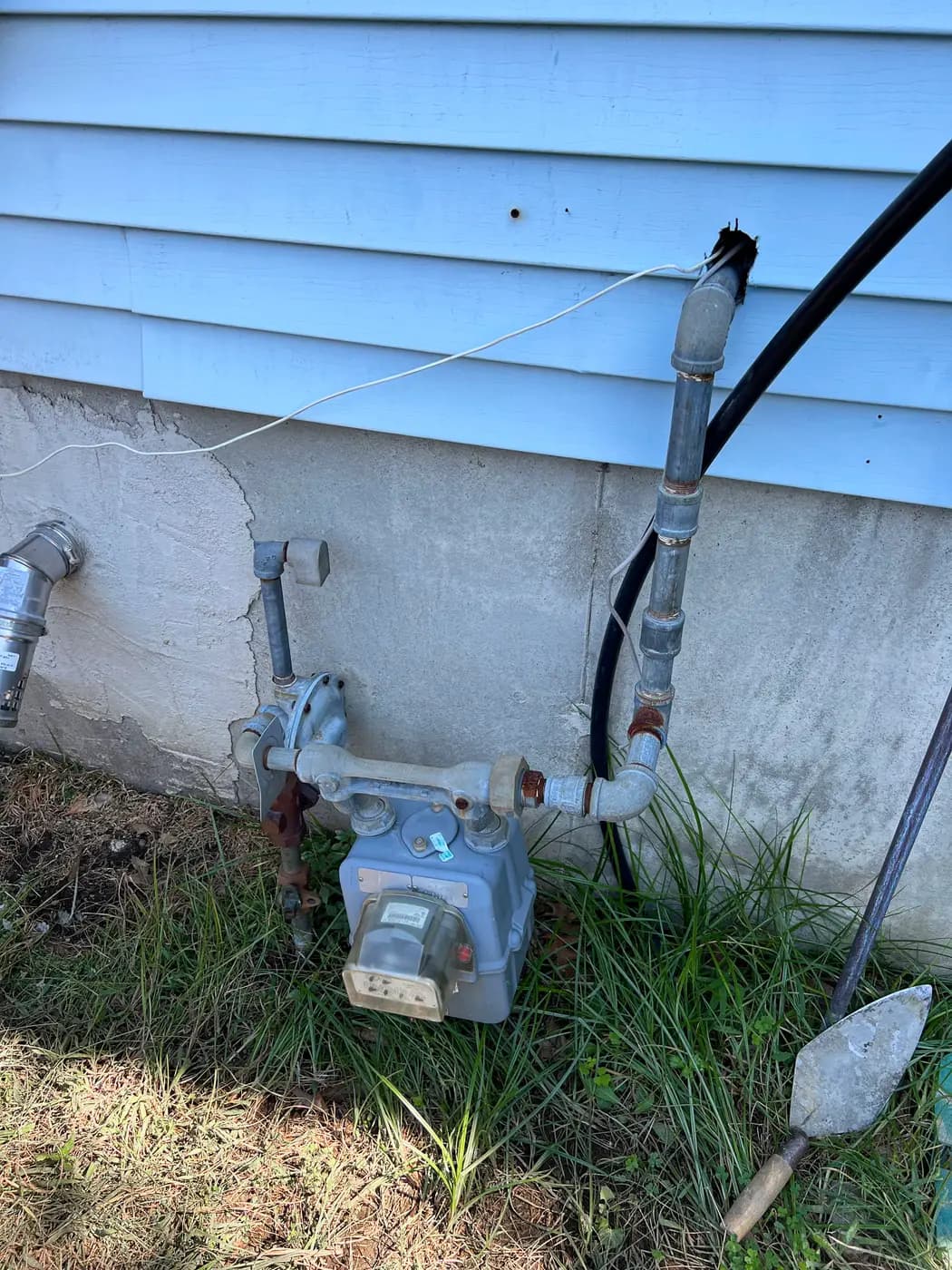 Gas meter and piping at blue sided house with shovel leaning nearby