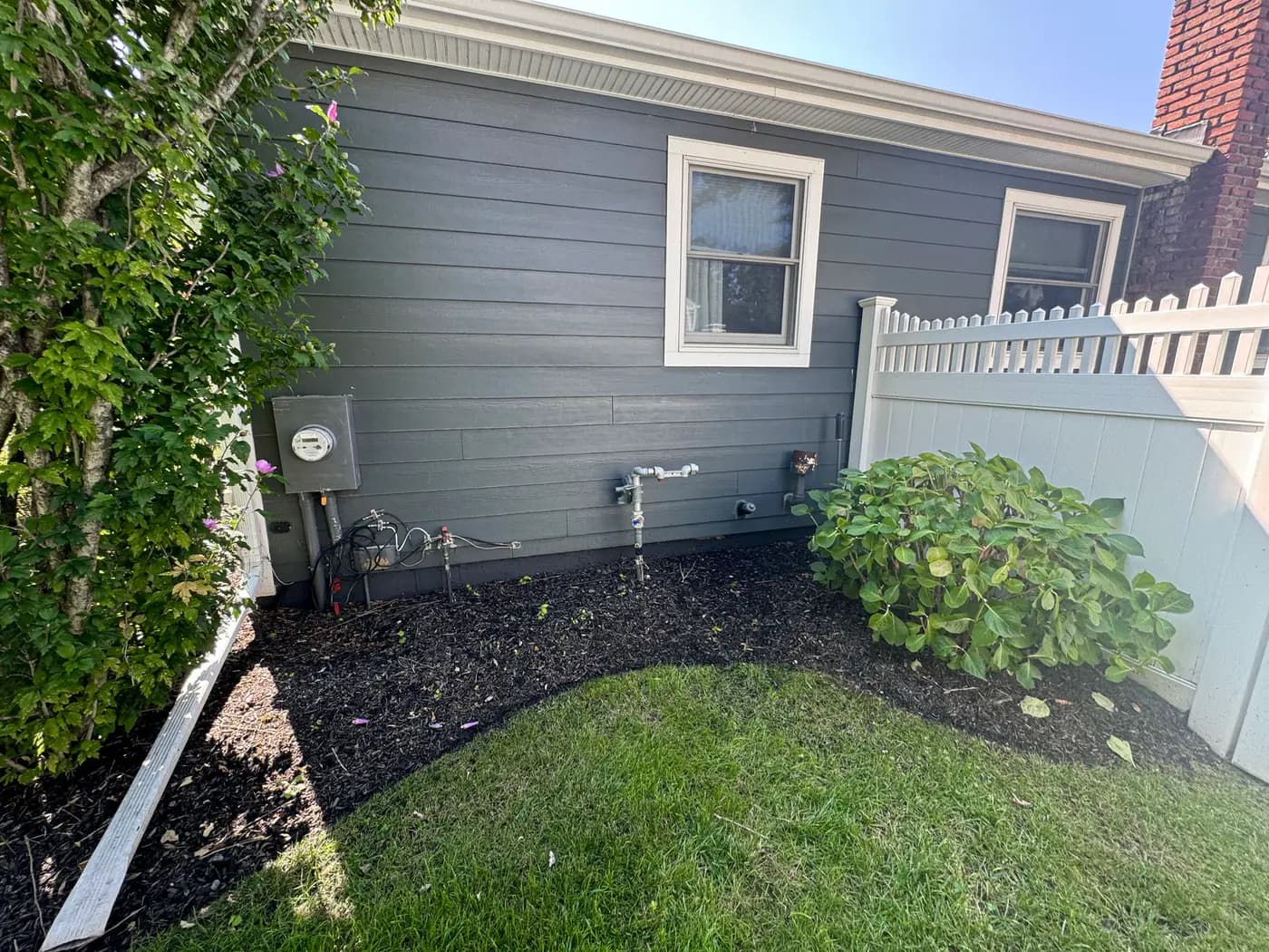 Rear corner of gray sided house with hose bib, mulch bed and lawn