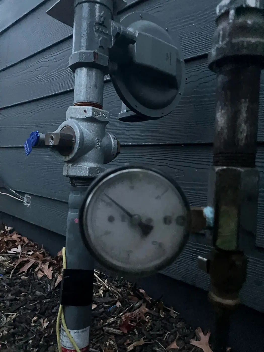 Gas regulator and pressure gauge on riser piping against dark gray siding