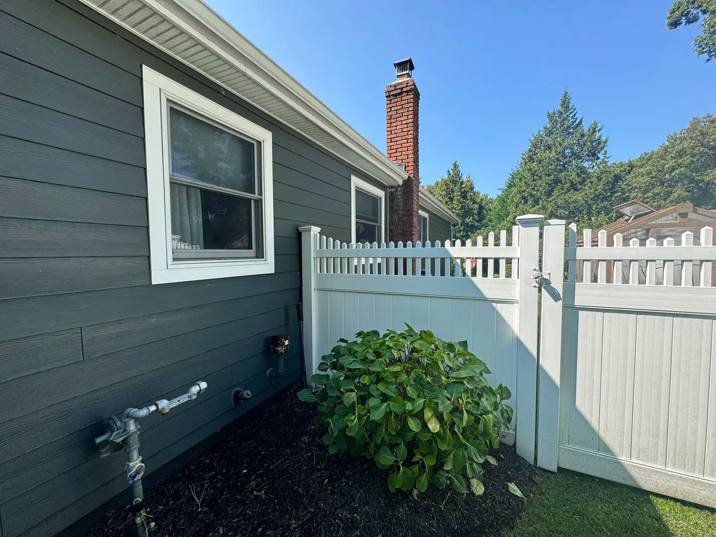 Exterior hose bib on gray siding beside shrub and white vinyl fence
